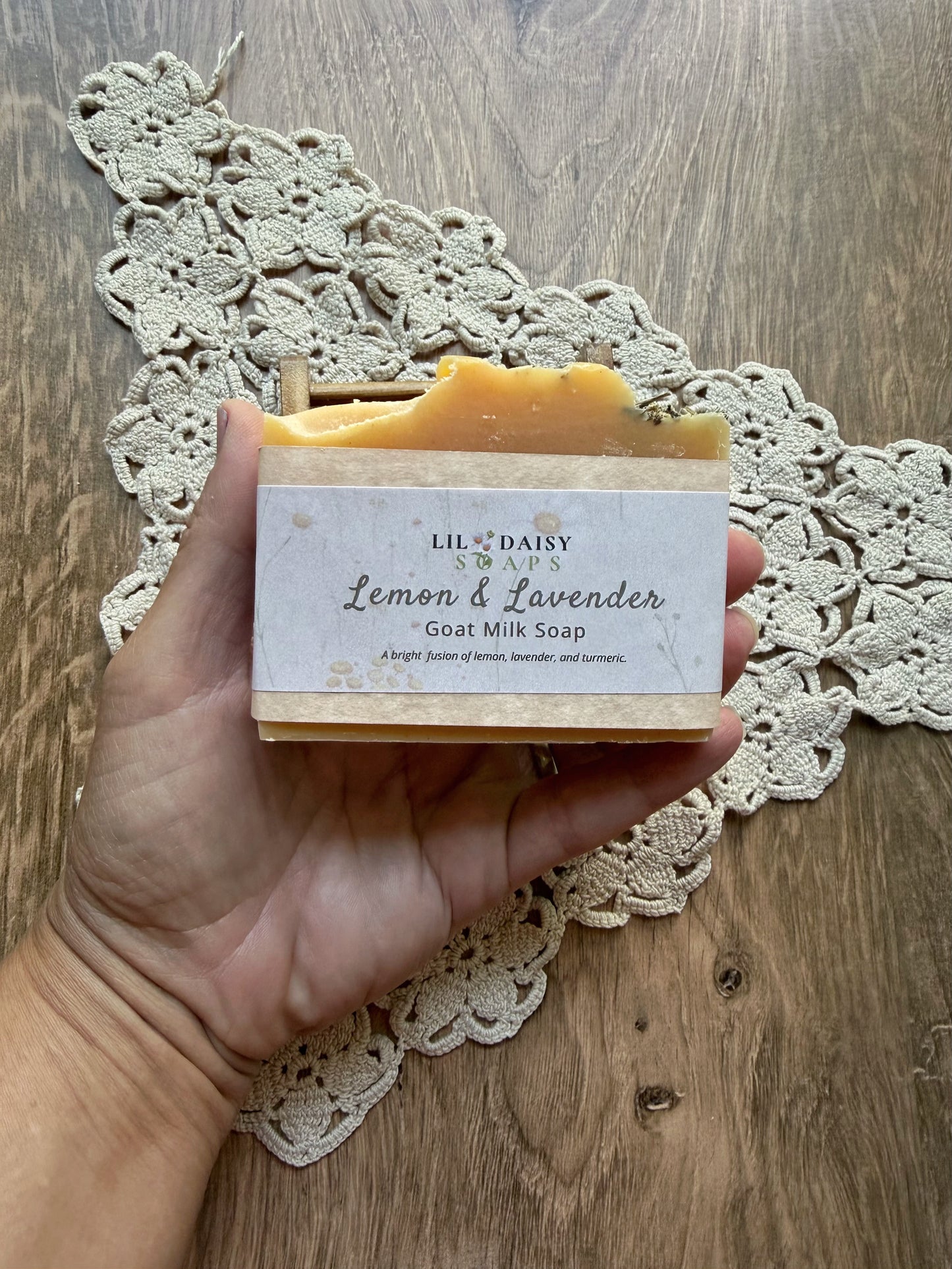 Hand holding Lil Daisy Soaps Lemon & Lavender Natural Goat Milk Soap Bar above a lace doily on wood.