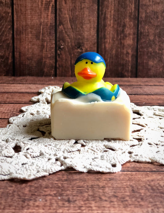 A rubber duck in a blue cap tops Lil Daisy Soaps Rubber Ducky Goat Milk Soap on a crocheted doily and wood.