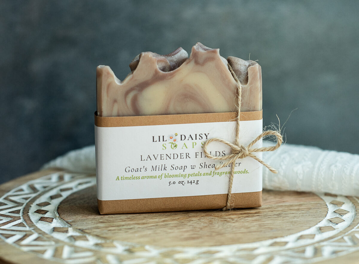 A handcrafted bar of marbled soap labeled Lil Daisy Soap: Lavender Fields sits on a wooden surface, wrapped in brown paper and twine, with a soft cloth and gray background behind it.