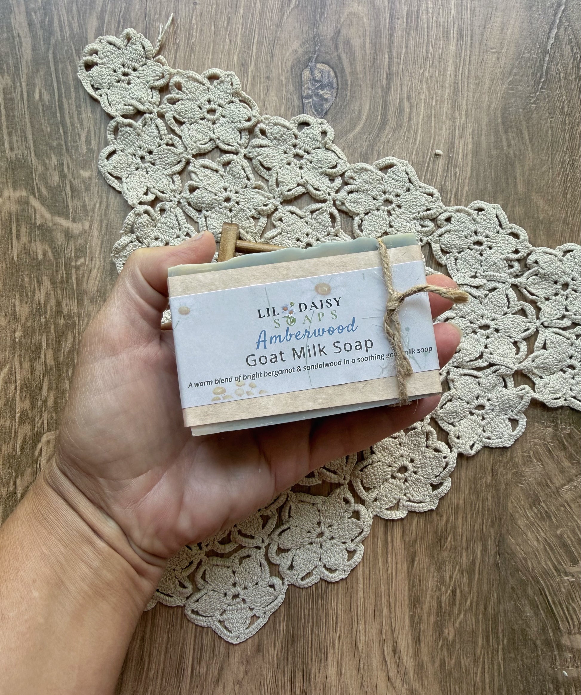 A hand holds Lil Daisy Soaps Amberwood Natural Goat Milk Soap Bar over a crocheted doily on wood.