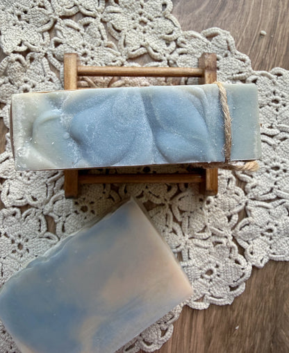 Amberwood Goat Milk Soap Bar by Lil Daisy Soaps on a wooden rack atop a crocheted lace mat.