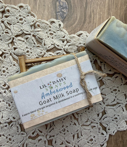 Amberwood Natural Goat Milk Soap Bar by Lil Daisy Soaps on a mat, with box and wooden soap holder nearby.