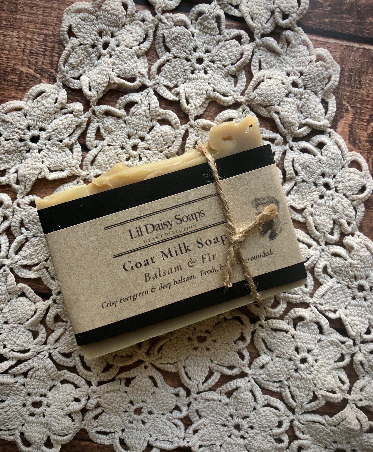 A bar of Lil Daisy Soaps Balsam & Fir Goat Milk Soap on a crocheted doily atop a wooden surface.