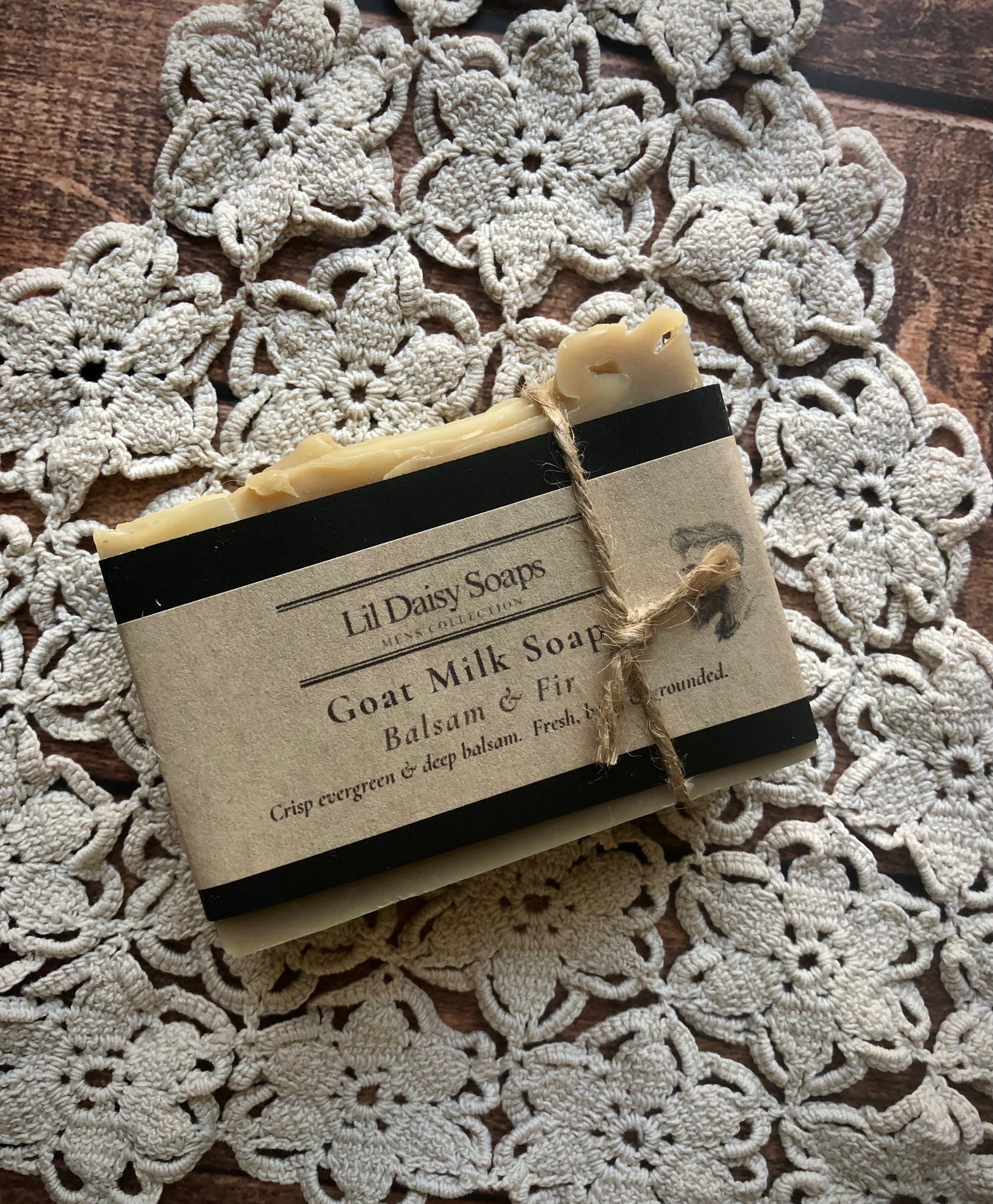 A bar of Lil Daisy Soaps Balsam & Fir Goat Milk Soap on a crocheted doily atop a wooden surface.