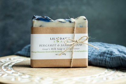 A bar of Lil Daisy Soaps Amberwood Natural Goat Milk Soap rests on a wooden tray next to a blue towel.