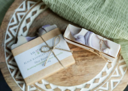 Two Lil Daisy Soaps bars, including Berries & Cream Goat Milk Soap, sit on a wooden tray with a green cloth behind.
