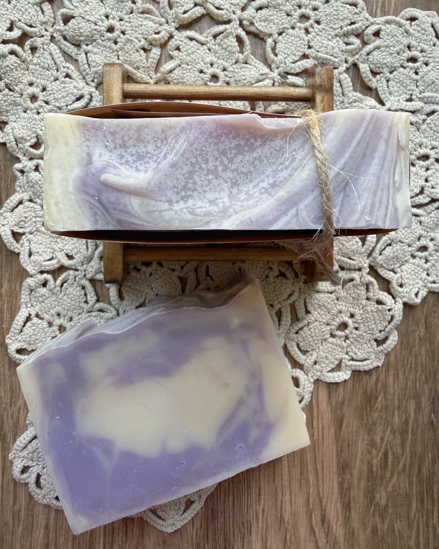 Two Lil Daisy Soaps Berry Creme Goat Milk Bars sit on a lace doily, one on a wooden soap dish.