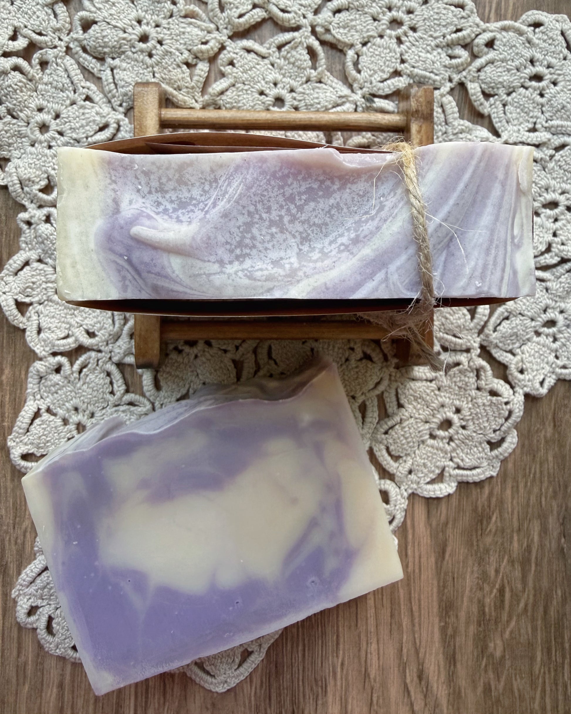 Two Lil Daisy Soaps Berry Creme Goat Milk Bars sit on a lace doily, one on a wooden soap dish.