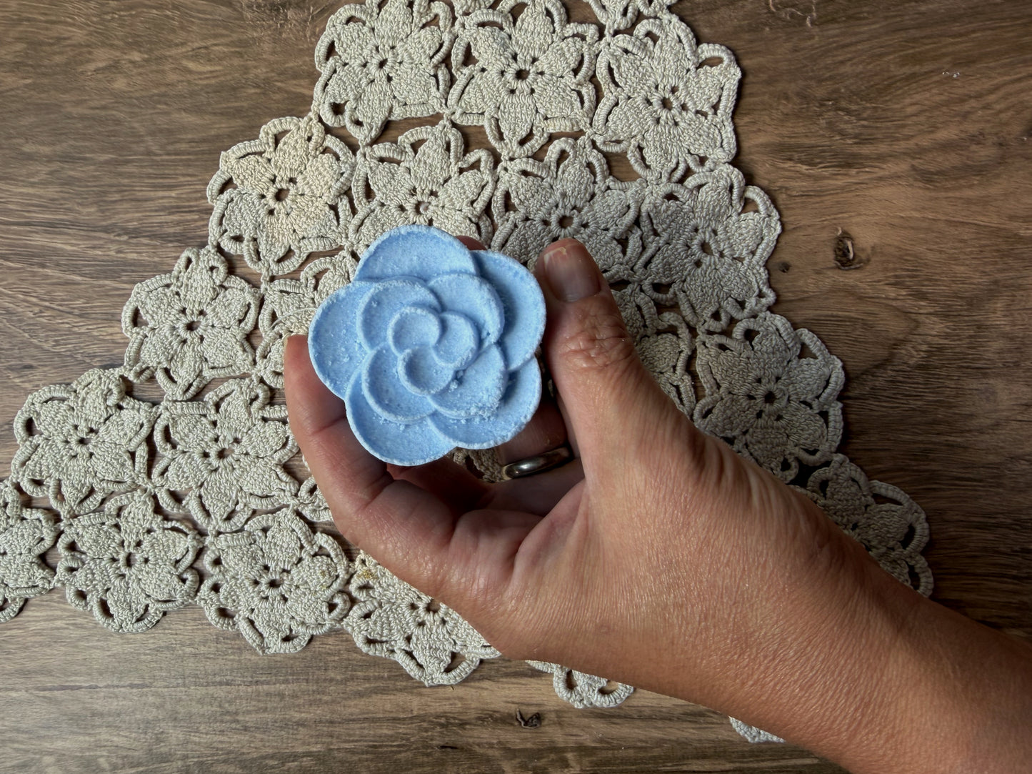 A hand holds a blue, flower-shaped Essential Oil Shower Steamer by Lil Daisy Soaps above a crocheted doily.