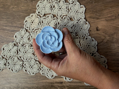 A hand holds a blue, flower-shaped Essential Oil Shower Steamer by Lil Daisy Soaps above a crocheted doily.