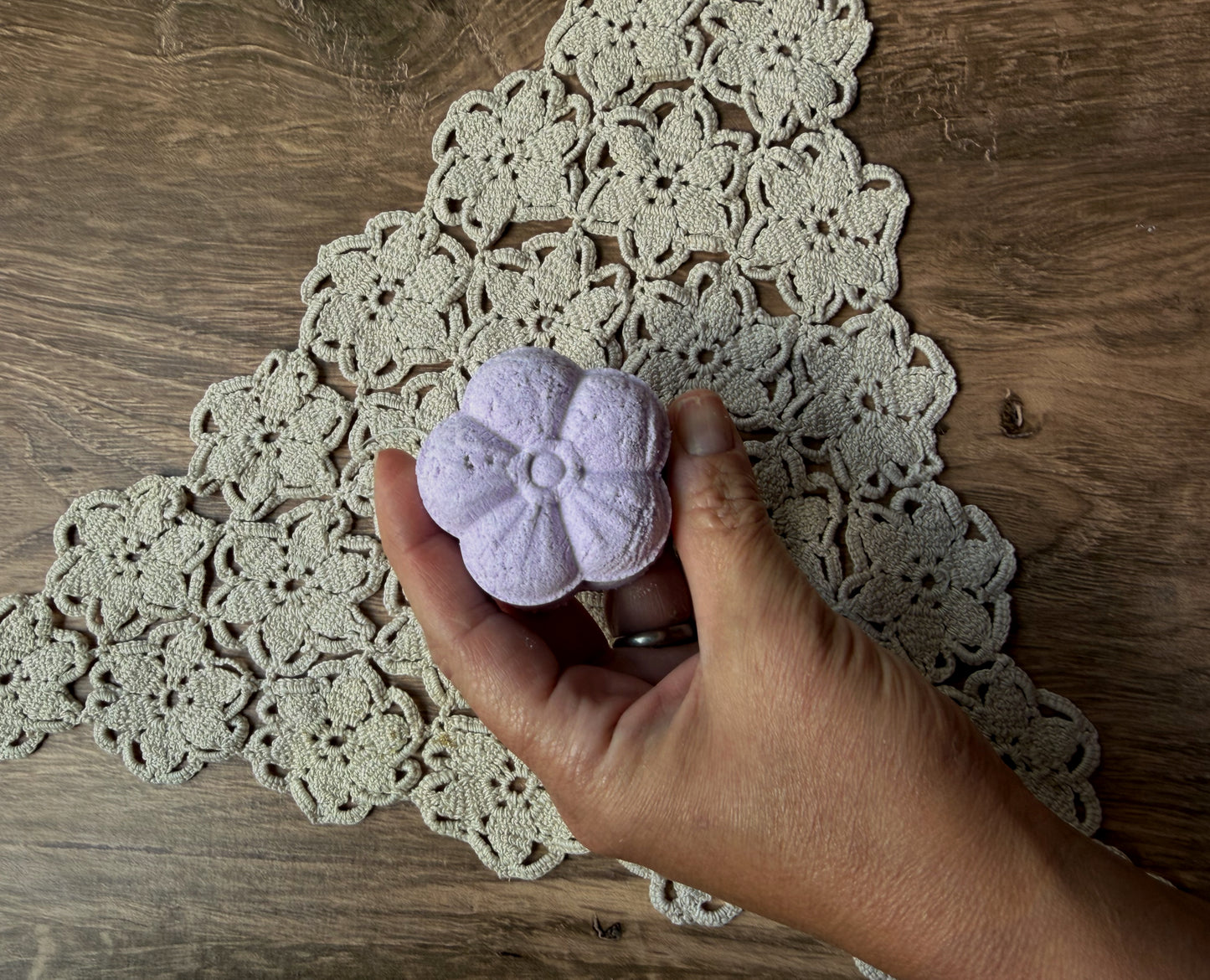 A hand holds a Lil Daisy Soaps Essential Oil Shower Steamer above a tan crocheted doily on a wooden table.