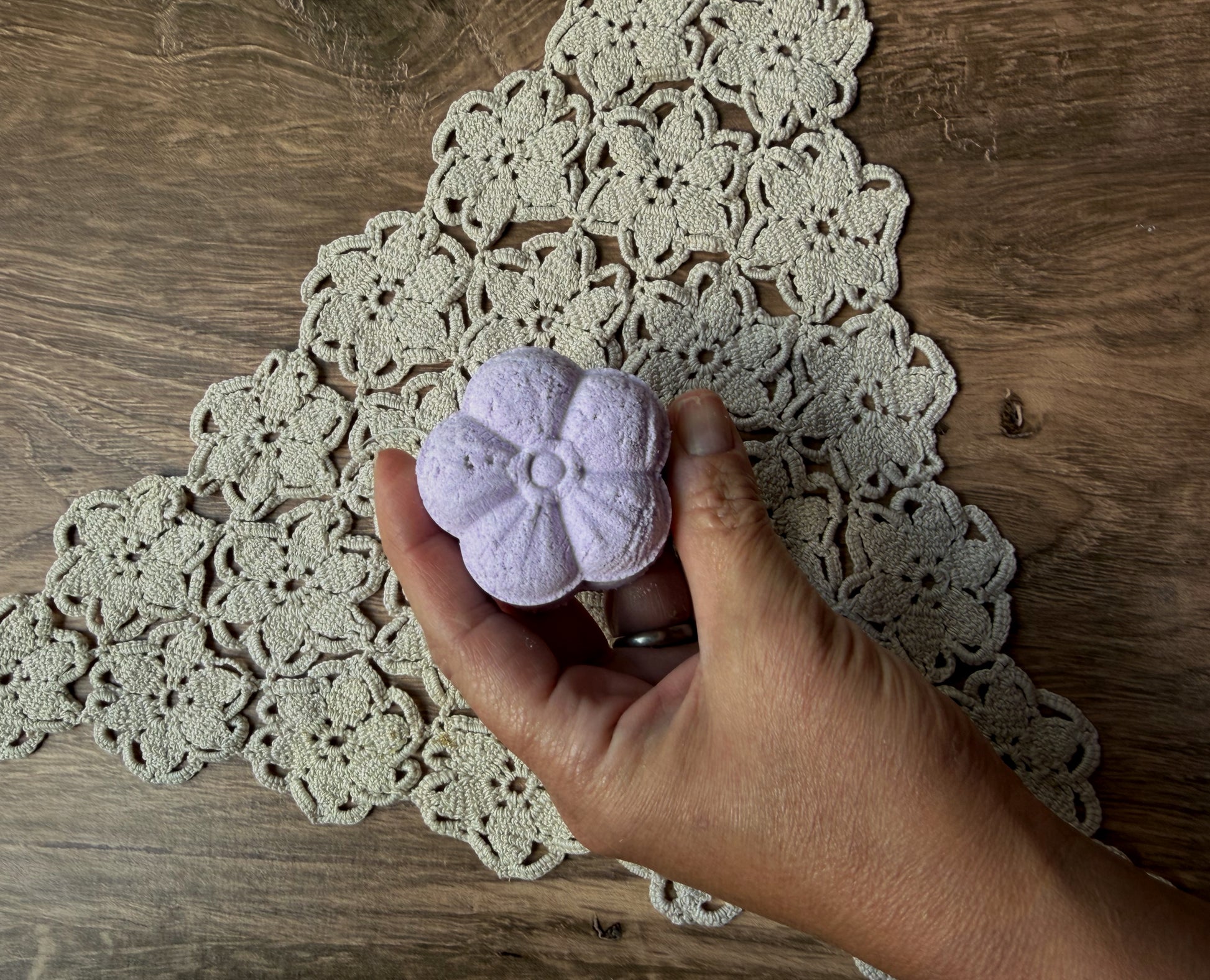 A hand holds a Lil Daisy Soaps Essential Oil Shower Steamer above a tan crocheted doily on a wooden table.