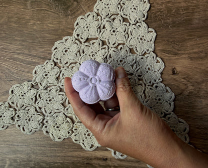 A hand holds a Lil Daisy Soaps Essential Oil Shower Steamer above a tan crocheted doily on a wooden table.