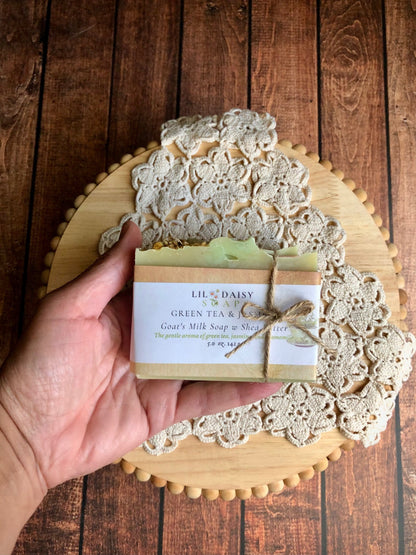 A hand holds Lil Daisy Soaps’ Green Tea & Jasmine Goat Milk Soap above a lace doily on a wooden board.