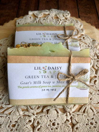 Two Lil Daisy Soaps Green Tea & Jasmine Goat Milk Soap bars sit wrapped in paper and twine on wood.
