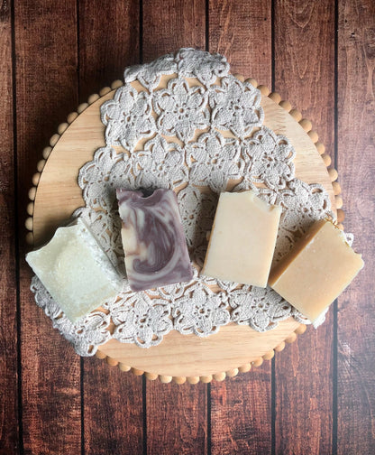 Four Lil Daisy Soaps Goat Milk Soap Sample Set bars on a lace doily, displayed on a wooden board.