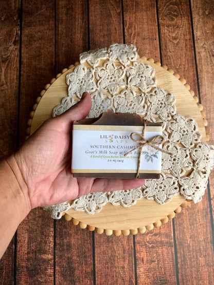 A hand holds Lil Daisy Soaps Southern Cashmere Goat Milk Soap above a wooden tray with a lace doily.