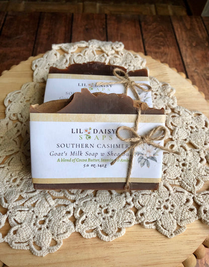 Two bars of Lil Daisy Soaps Southern Cashmere Goat Milk Soap sit on a crocheted doily, wrapped in paper.