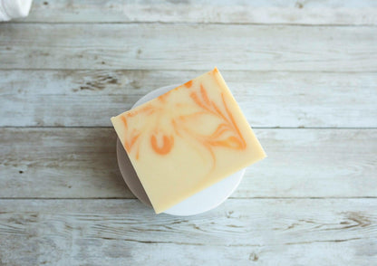 A Lil Daisy Soaps Orange Patchouli Natural Goat Milk Soap Bar with orange swirls rests on a white dish.