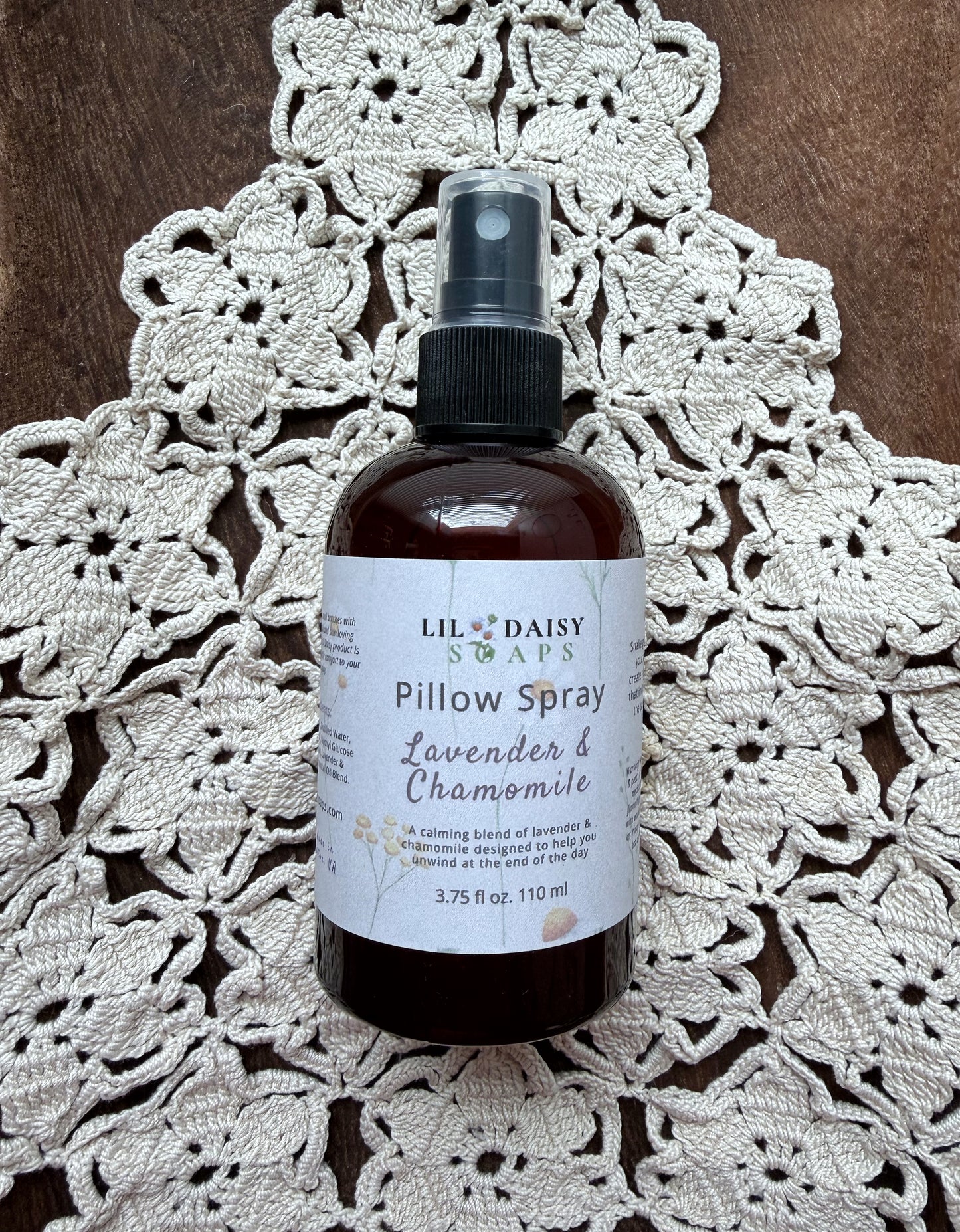 Lil Daisy Soaps’ Lavender & Chamomile Pillow Spray sits on a crocheted doily atop a wooden surface.