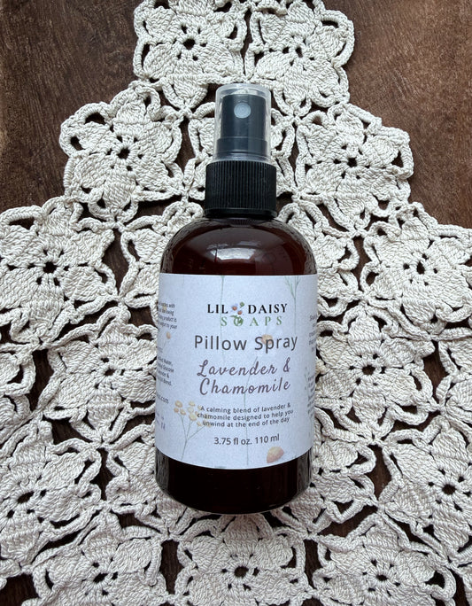 Lil Daisy Soaps’ Lavender & Chamomile Pillow Spray sits on a crocheted doily atop a wooden surface.