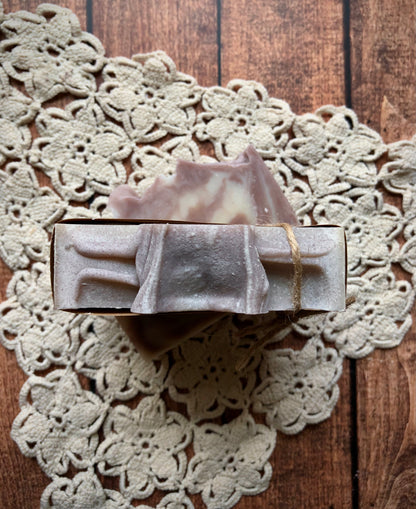 The Lavender Fields Natural Goat Milk Soap Bar by Lil Daisy Soaps sits on a doily, tied with twine.