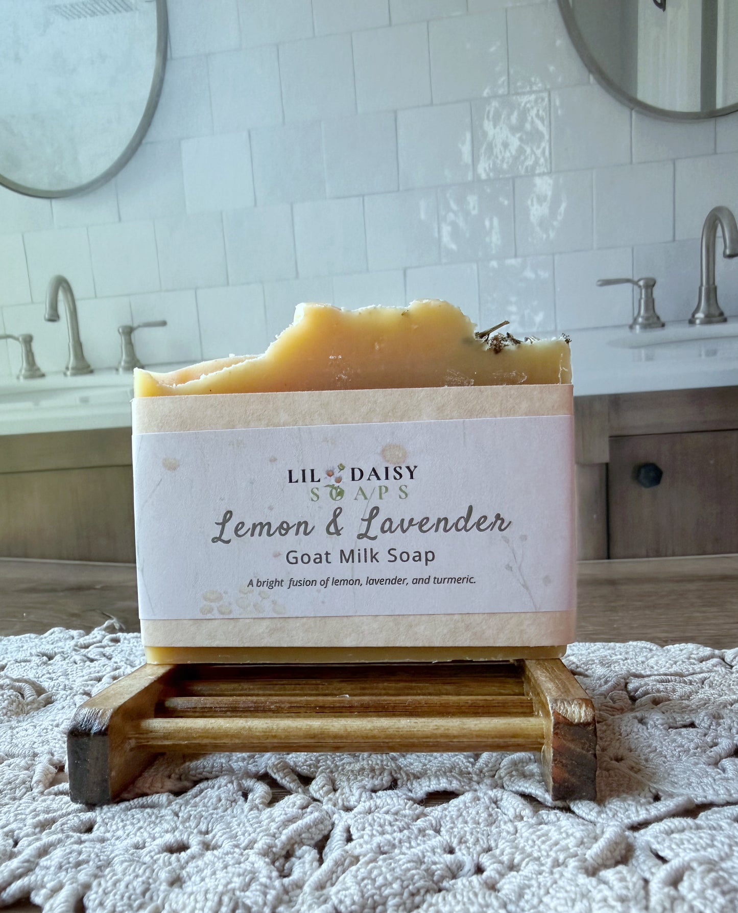 Lil Daisy Soaps’ Lemon & Lavender Goat Milk Soap Bar sits on a wooden holder in a bathroom with sinks and mirrors.