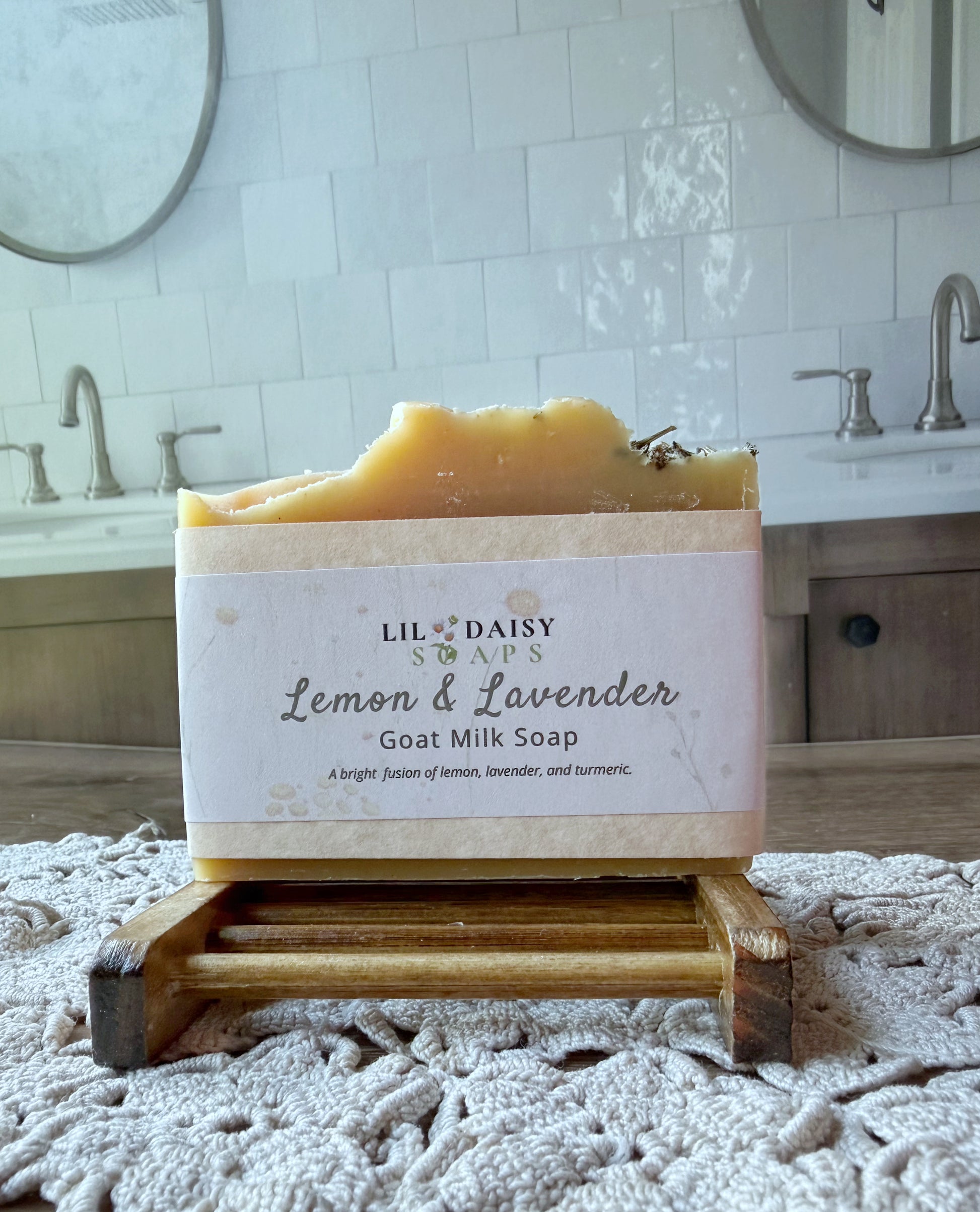 Lil Daisy Soaps’ Lemon & Lavender Goat Milk Soap Bar sits on a wooden holder in a bathroom with sinks and mirrors.