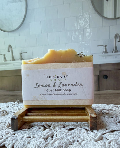 Lil Daisy Soaps’ Lemon & Lavender Goat Milk Soap Bar sits on a wooden holder in a bathroom with sinks and mirrors.