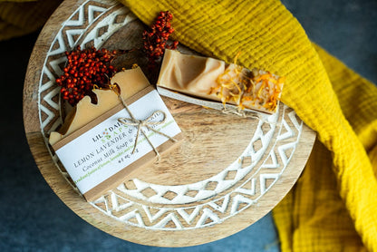 Two Lil Daisy Soaps Lemon & Lavender Goat Milk bars on a wooden tray with a yellow cloth and red flowers.