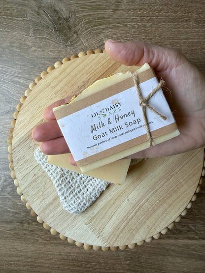 Hand holds Lil Daisy Soaps’ Milk & Honey Goat Milk Soap Bar with oats, above a wood board and soap saver bag.