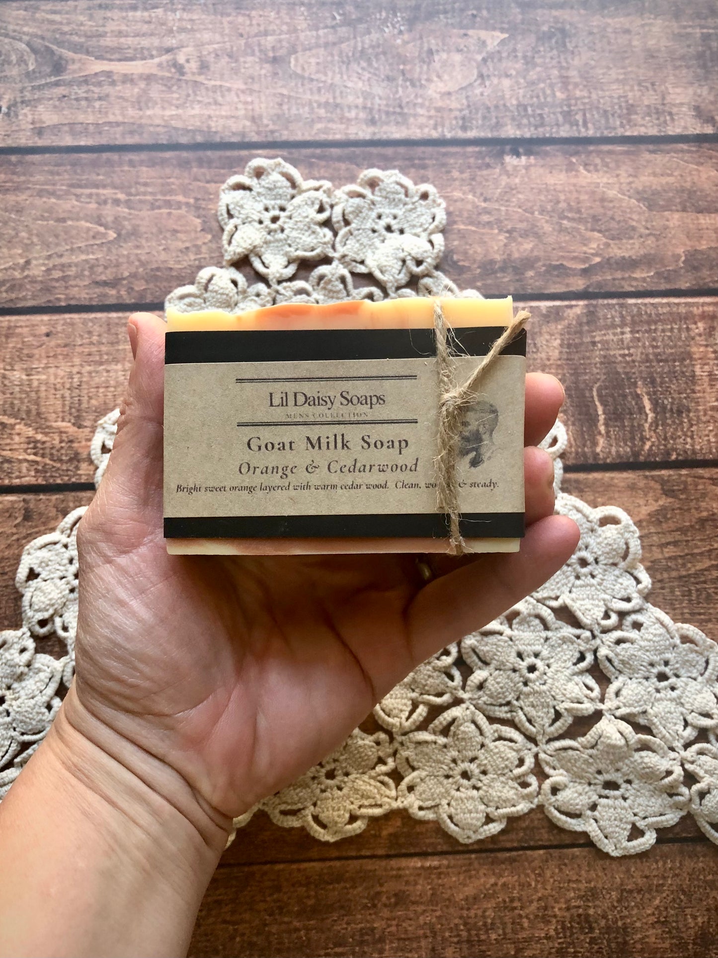 Hand holding Lil Daisy Soaps Orange & Cedarwood Natural Goat Milk Soap over lace and wood background.