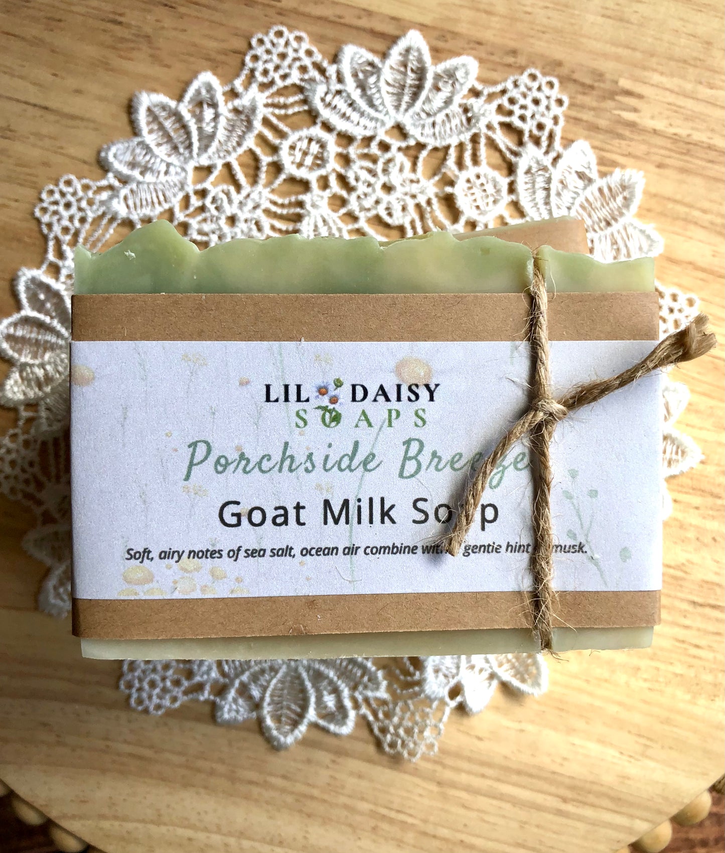 A bar of Porchside Breeze Goat Milk Soap by Lil Daisy Soaps rests on a lace doily atop a wooden surface.