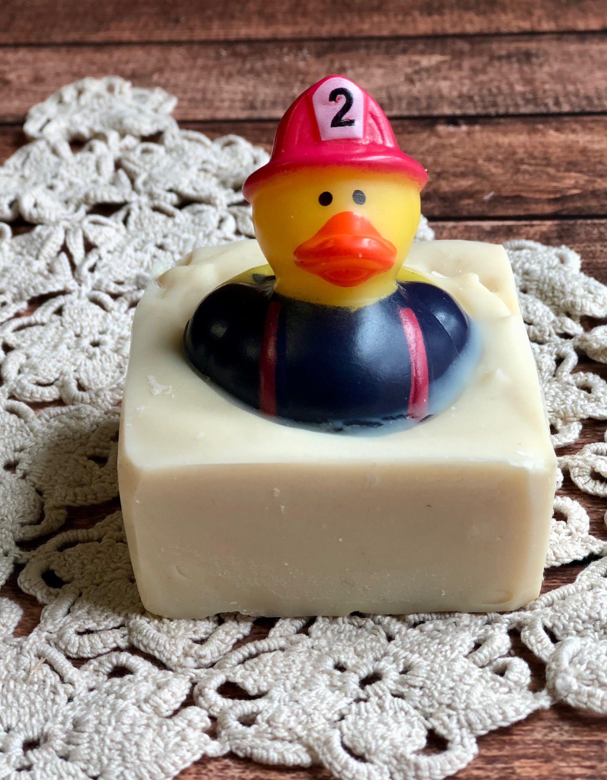 A firefighter-hat rubber duck sits atop Lil Daisy Soaps’ Rubber Ducky Goat Milk Soap on a crocheted doily.