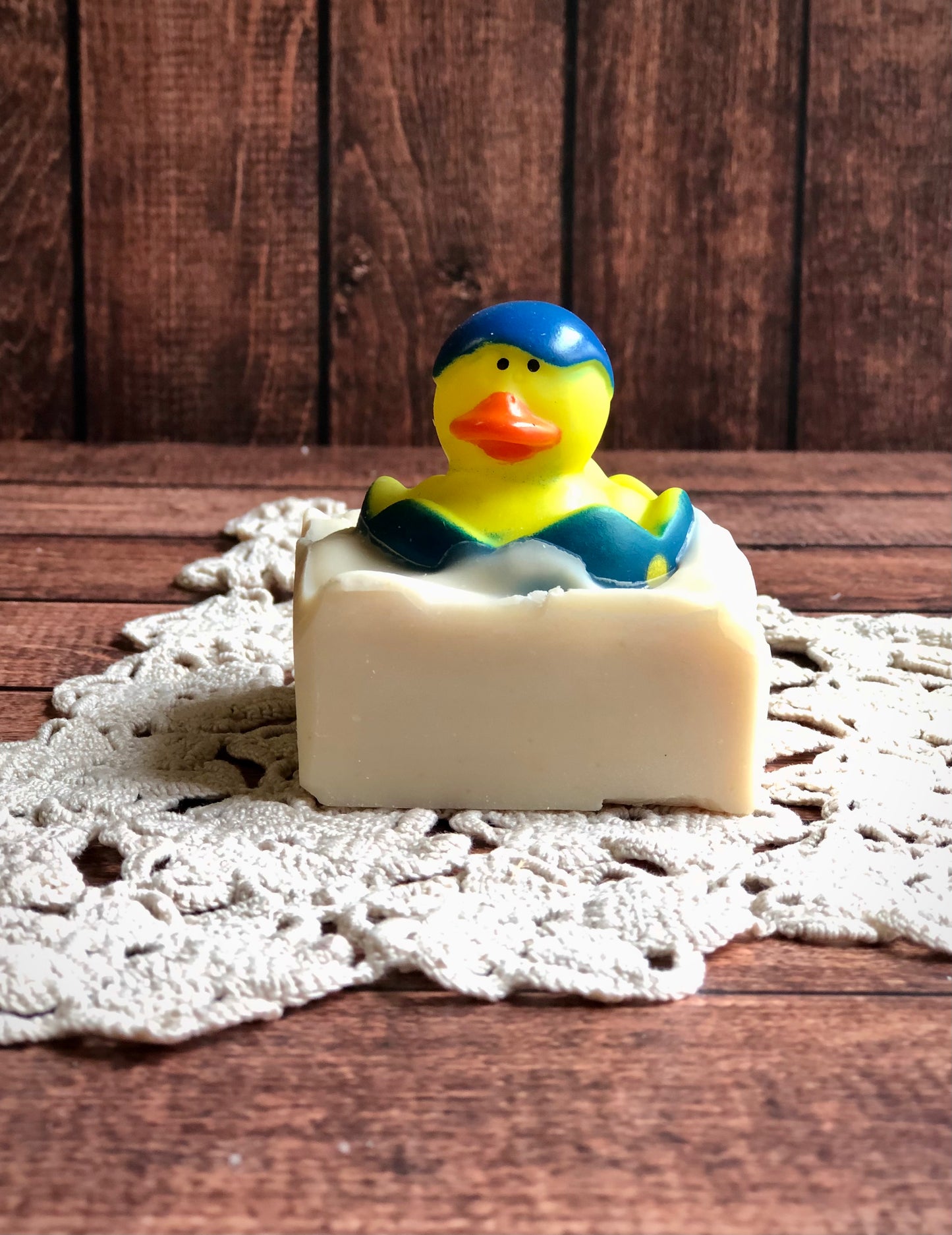 A rubber duck in a blue cap tops Lil Daisy Soaps Rubber Ducky Goat Milk Soap on a crocheted doily and wood.