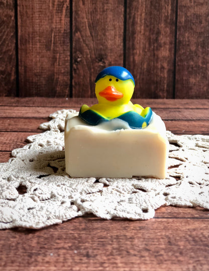 A rubber duck in a blue cap tops Lil Daisy Soaps Rubber Ducky Goat Milk Soap on a crocheted doily and wood.