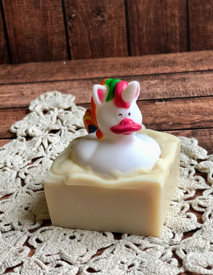 A unicorn rubber duck sits atop Lil Daisy Soaps Rubber Ducky Goat Milk Soap on a lace doily and wood surface.