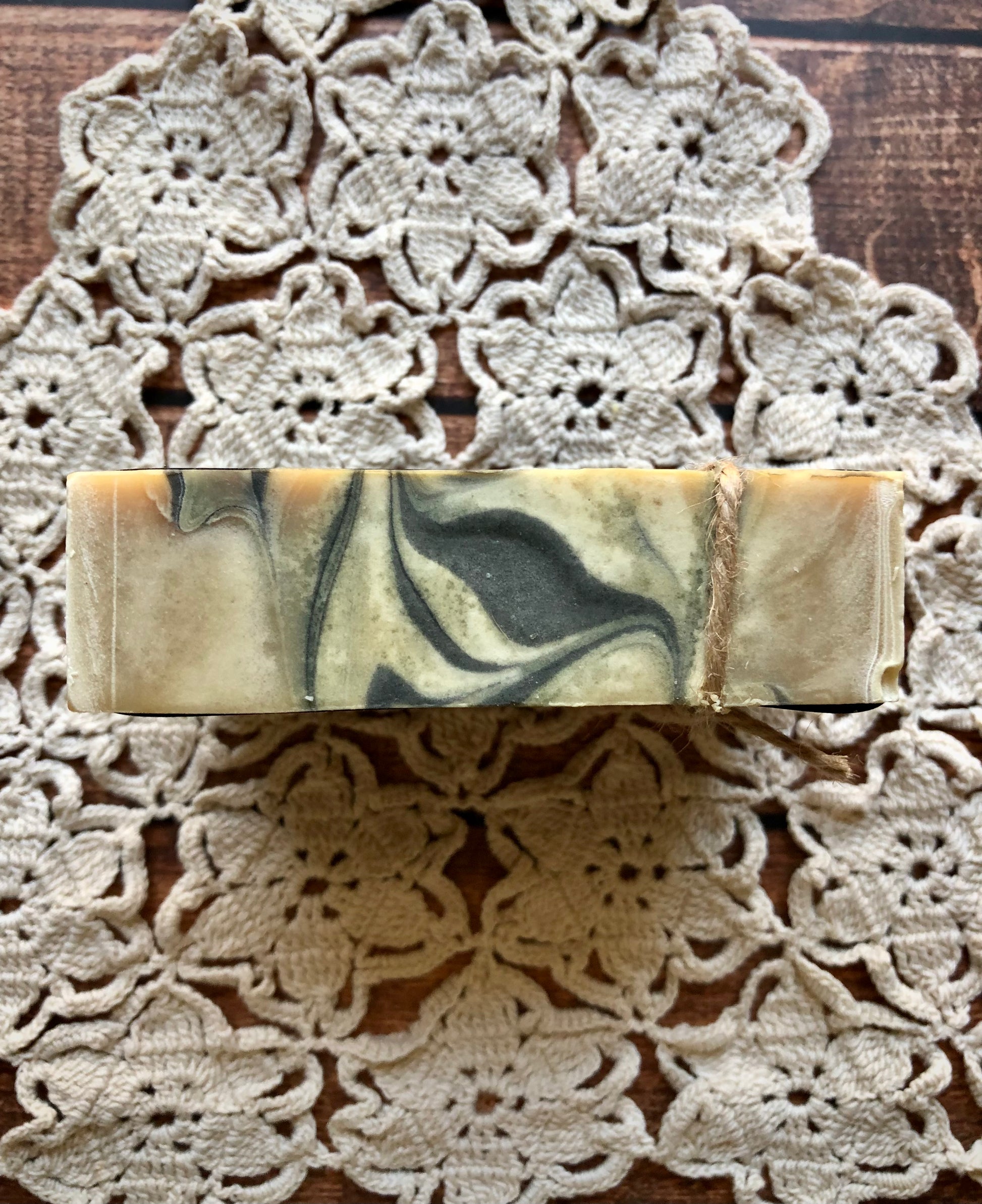 Lil Daisy Soaps Sandalwood & Musk Goat Milk & Beer Soap with twine sits on a floral doily on wood.