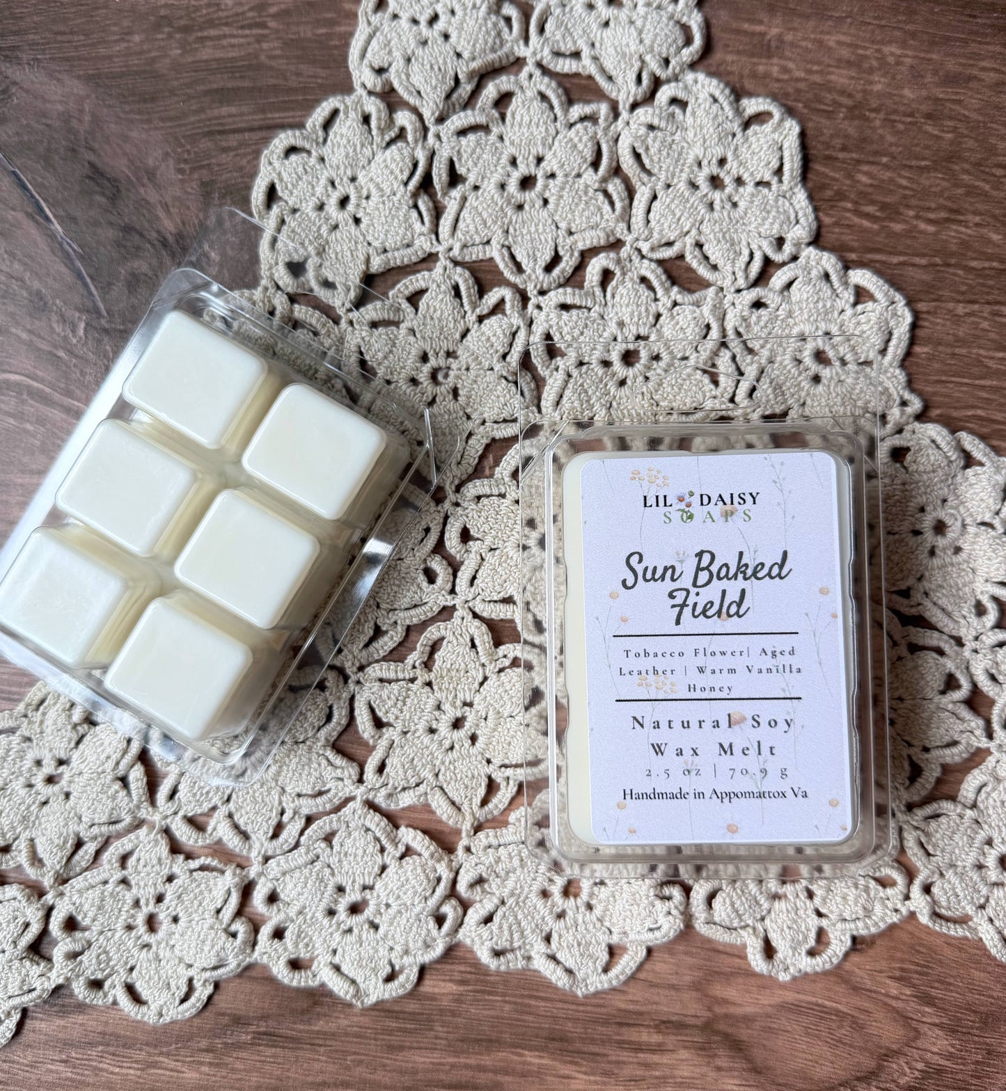 Two Lil Daisy Soaps wax melts, one open, one Sun Baked Field, rest on a doily over wood.