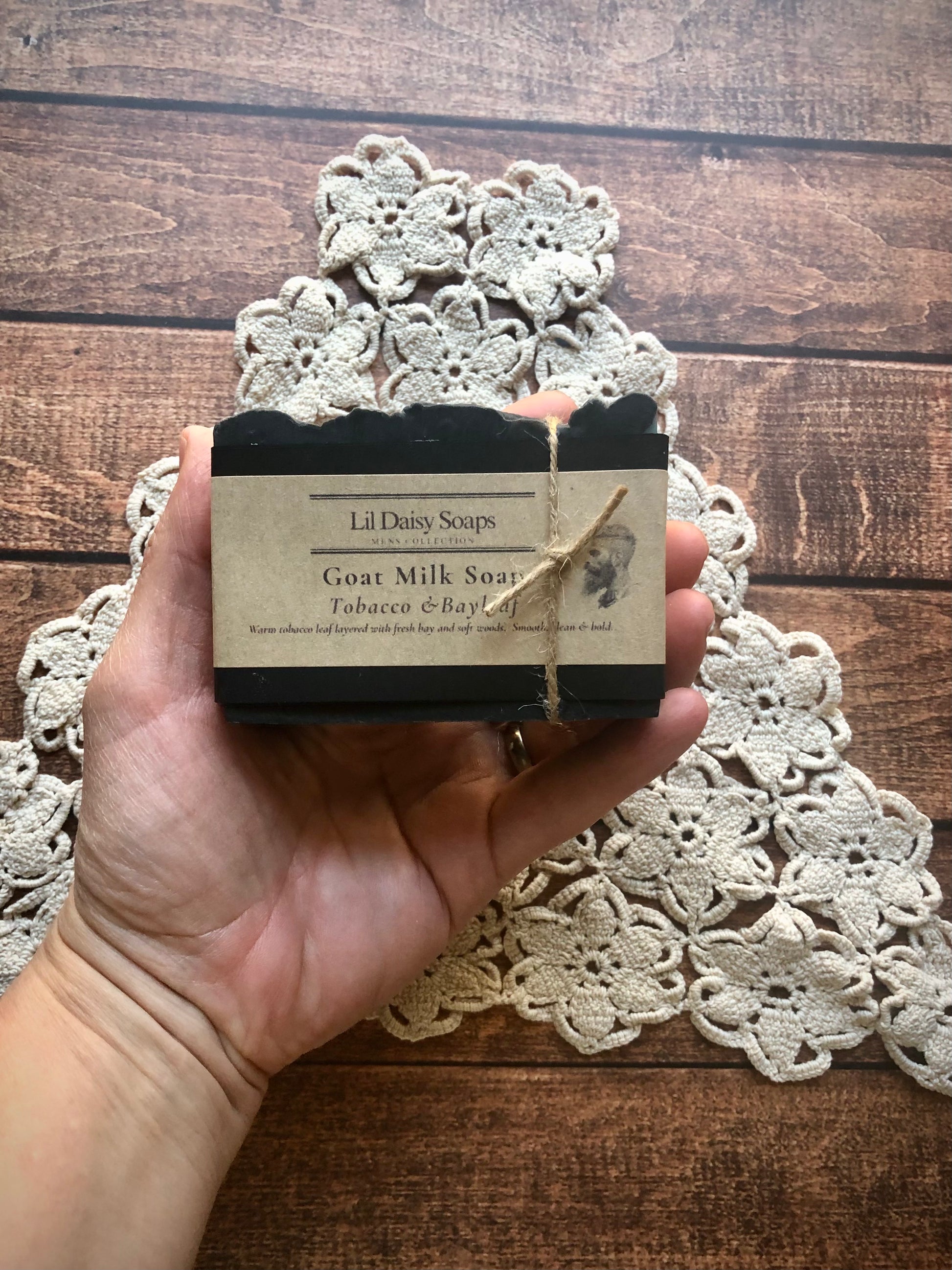 A hand holds Lil Daisy Soaps Tobacco & Bayleaf Goat Milk Soap Bar on a crocheted doily atop wood.